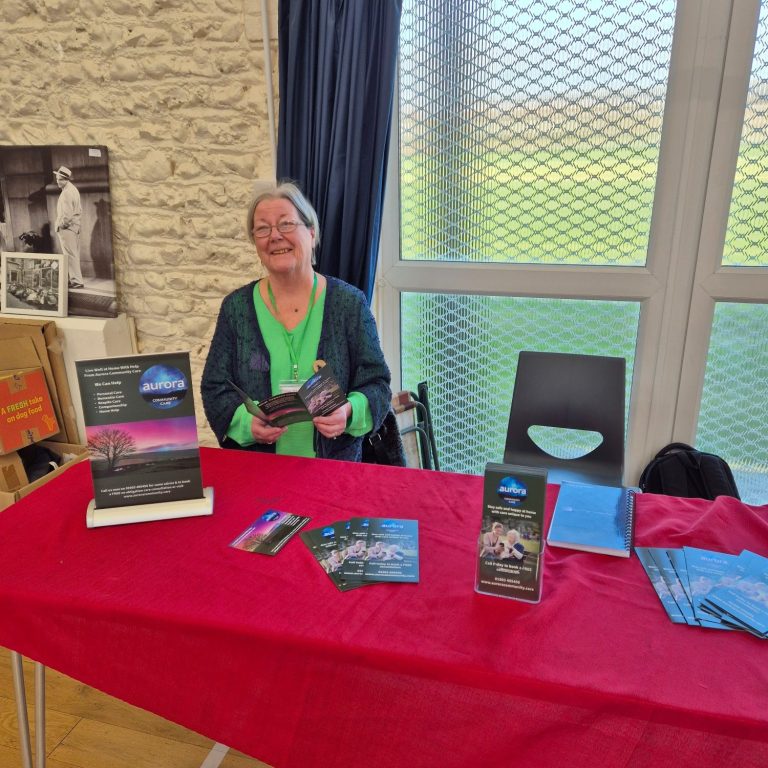A smiling woman sits at a table with brochures and a book in a well-lit room.