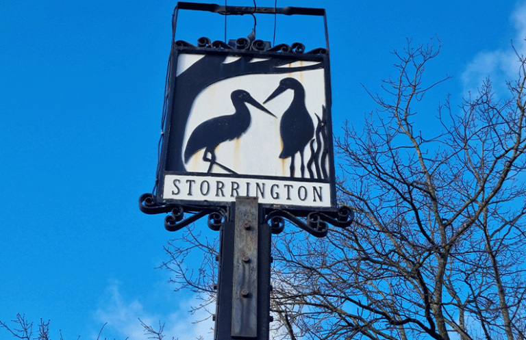 Sign for Storrington featuring two storks on a white background against a blue sky.