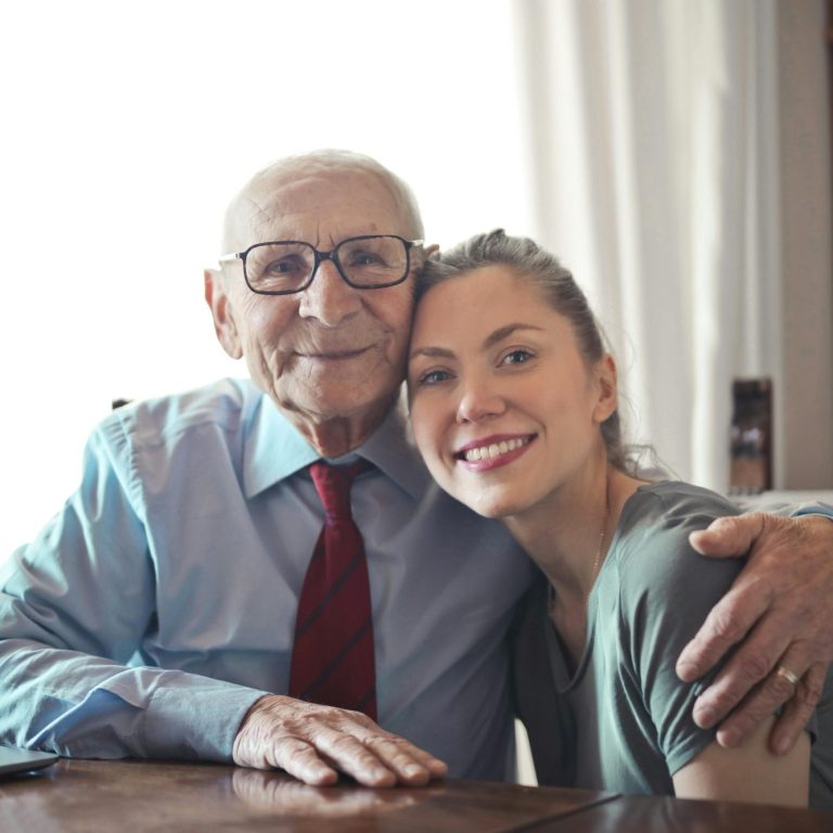 An elderly man and a young woman smiling together, sitting closely at a table.