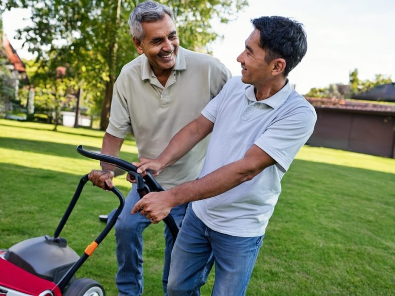 Two men smiling while pushing a lawnmower on a grassy area.