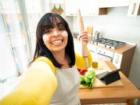 Smiling Home Care Giver in a kitchen holding a rolling pin, surrounded by vegetables and a tablet.