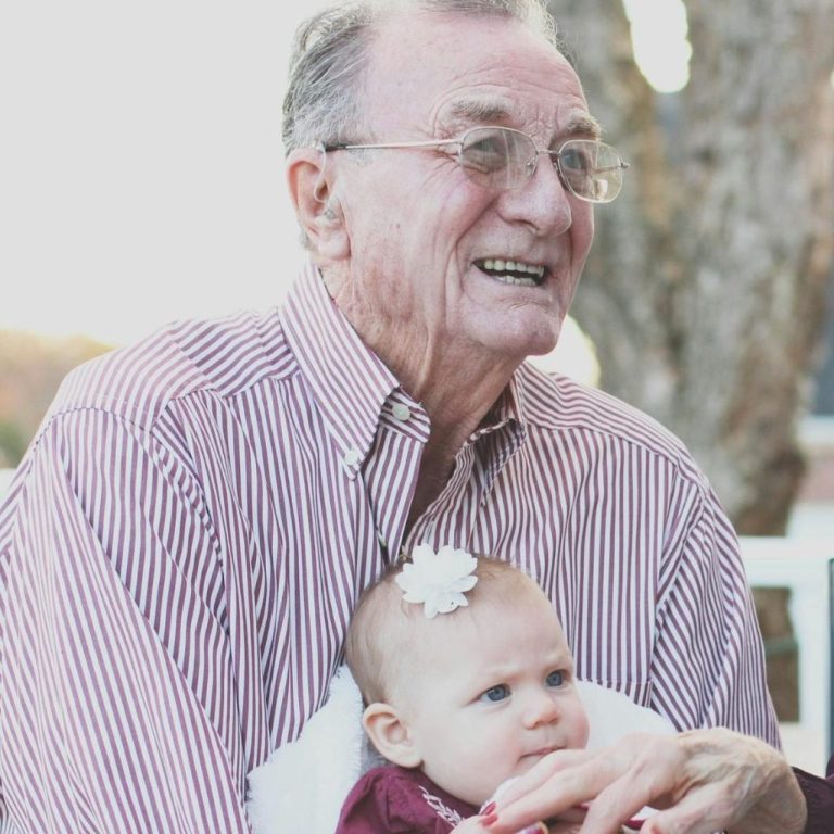 An elderly man smiling while holding a baby girl with a flower headband.
